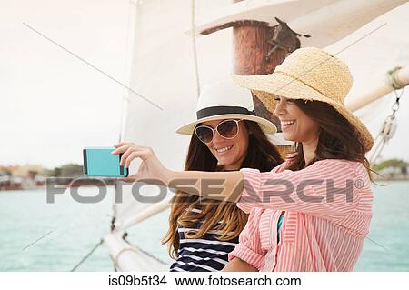 Picture - Two women on sailing boat, taking selfie, using smartphone. Fotosearch