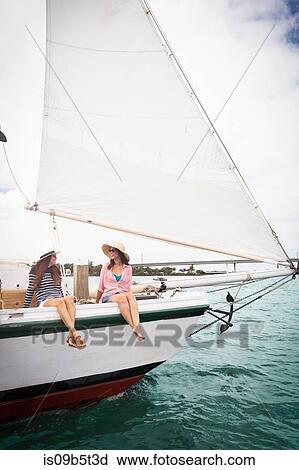 Stock Image - Two women on sitting on edge of sailing boat, legs dangling over the edge. Fotosearch