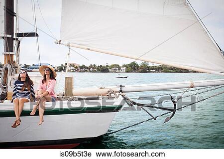 Stock Image - Two women on sitting on edge of sailing boat, legs dangling over the edge. Fotosearch