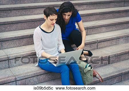 Stock Image - Two women, sitting on steps, looking at laptop. Fotosearch