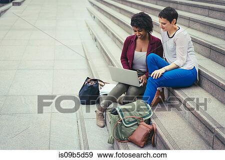 Stock Photo - Two women, sitting on steps, looking at laptop. Fotosearch