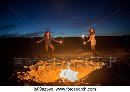Stock Image - Two young girls holding sparklers on beach beside campfire, holding sparklers. Fotosearch