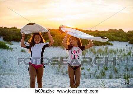 Stock Image - Two young girls on beach, carrying surfboards. Fotosearch