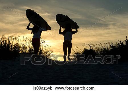 Stock Image - Two young girls on sand dunes, at dusk, carrying surfboards. Fotosearch