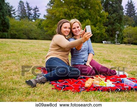 Stock Image - Two young women enjoying picnic in park, taking selfie, using smartphone. Fotosearch