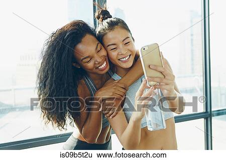 Two young women in gym, taking selfie, using smartphone View Large Photo Image Stock Photograph - Two young women in gym, taking selfie, using smartphone. Fotosearch