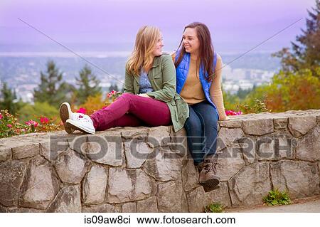 Two young women, sitting on wall, face to face, smiling View Large Photo Image Stock Image - Two young women, sitting on wall, face to face, smiling. Fotosearch