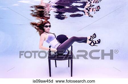 Stock Image - Underwater side view of mid adult woman wearing high heels and sunglasses in swimming pool sitting on chair looking at camera. Fotosearch