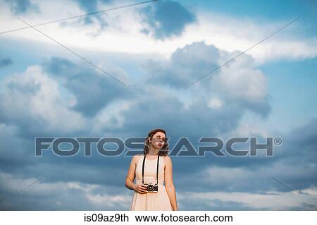 Stock Image - Woman against cloudy blue sky holding camera. Fotosearch