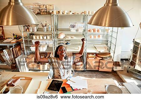 Stock Photograph - Woman at desk in pottery studio with paperwork, arms raised smiling. Fotosearch