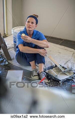 Stock Image - Woman decorating, taking a break, sitting on floor holding apple. Fotosearch