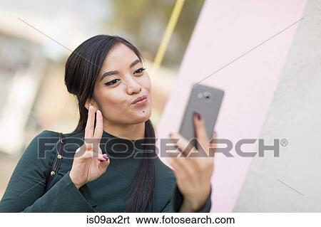 Stock Photograph - Woman doing peace sign and pouting using smartphone to take selfie. Fotosearch