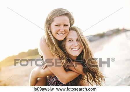 Stock Photo - Woman on beach giving friend piggyback looking at camera smiling. Fotosearch