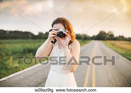 Stock Image - Woman on rural road taking photograph using camera. Fotosearch