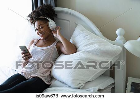 Stock Photo - Woman relaxing on bed, wearing headphones, looking at smartphone. Fotosearch