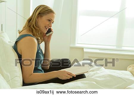 Stock Image - Woman sitting on bed chatting on smartphone. Fotosearch