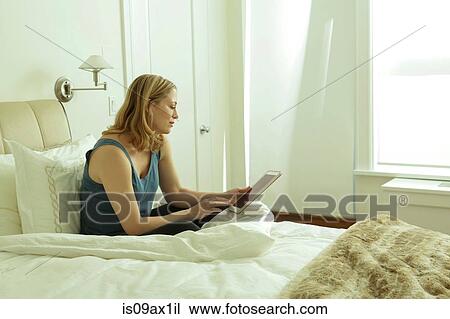 Stock Image - Woman sitting on bed reading digital tablet. Fotosearch