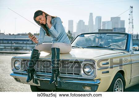 Stock Image - Woman sitting on convertible car bonnet looking at smartphone, Los Angeles, California, USA. Fotosearch