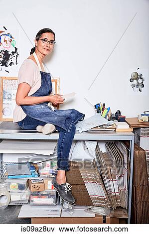 Woman sitting on desk wearing apron looking at camera smiling View Large Photo Image Stock Photo - Woman sitting on desk wearing apron looking at camera smiling. Fotosearch
