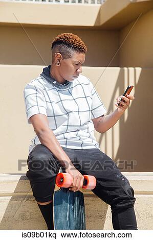 Woman sitting on wall holding skateboard looking at smartphone View Large Photo Image Stock Photography - Woman sitting on wall holding skateboard looking at smartphone. Fotosearch