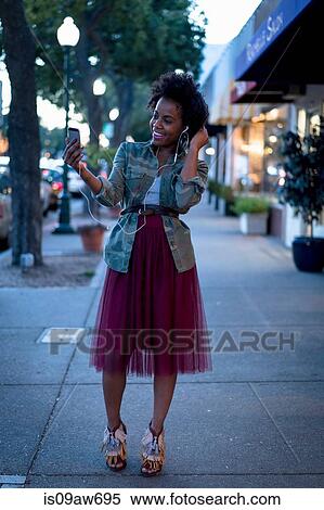 Woman standing in street, holding smartphone, wearing earphones, smiling View Large Photo Image Stock Photography - Woman standing in street, holding smartphone, wearing earphones, smiling. Fotosearch