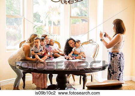 Stock Image - Woman taking photograph of mothers and babies at dining table. Fotosearch