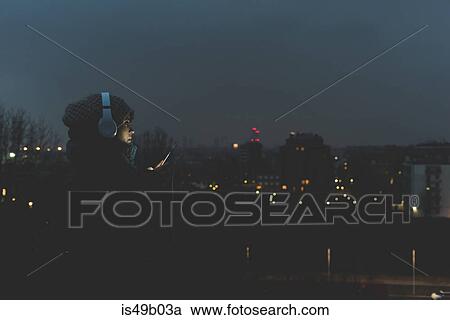 Woman wearing headphones looking at smartphone in city at night View Large Photo Image Stock Image - Woman wearing headphones looking at smartphone in city at night. Fotosearch