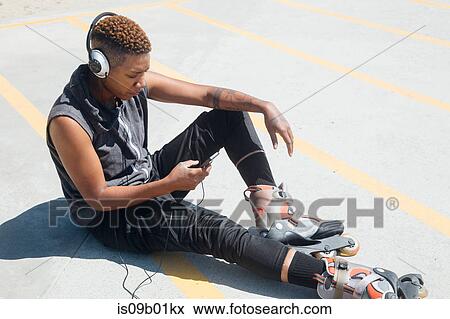 Stock Photo - Woman wearing rollerblades and headphones sitting on floor looking at smartphone. Fotosearch