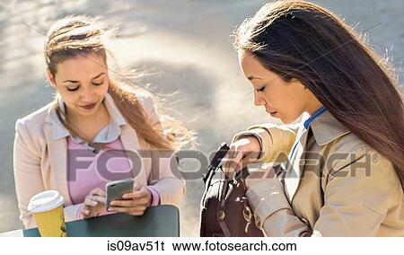 Stock Photograph - Young adult female twins reading smartphone texts at sidewalk cafe. Fotosearch