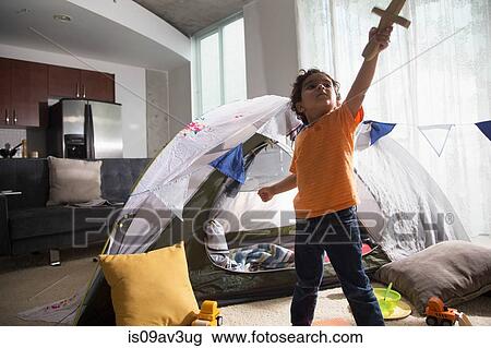 Stock Image - Young boy in living room using tent as den, arm raised, looking up, holding sword. Fotosearch