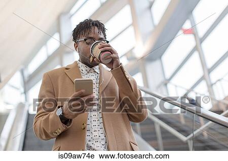 Stock Image - Young businessman moving down train station escalator drinking takeaway coffee. Fotosearch