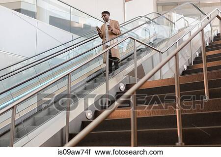 Stock Image - Young businessman moving down train station escalator with takeaway coffee. Fotosearch