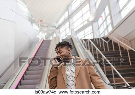 Stock Image - Young businessman talking on smartphone moving down train station escalator. Fotosearch