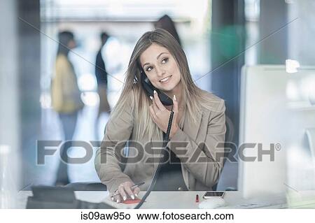 Young businesswoman sitting at desk, using telephone, smiling View Large Photo Image Picture - Young businesswoman sitting at desk, using telephone, smiling. Fotosearch