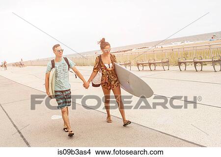 Picture - Young couple holding hands and carrying surfboards at Rockaway Beach, New York State, USA. Fotosearch