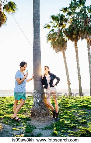 Stock Image - Young couple leaning against sunlit palm tree eating ice cream cones, Venice Beach, California, USA. Fotosearch