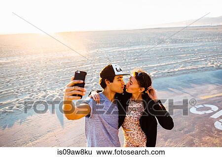 Stock Photograph - Young couple puckering up for smartphone selfie on beach, Venice Beach, California, USA. Fotosearch