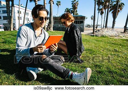 Young couple sitting on grass beside beach, woman wearing earphones, man looking at digital tablet View Large Photo Image Stock Image - Young couple sitting on grass beside beach, woman wearing earphones, man looking at digital tablet. Fotosearch