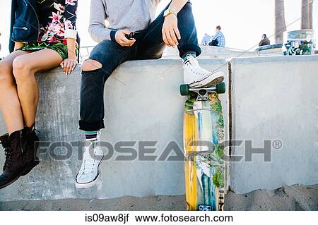 Stock Image - Young couple sitting on wall, man resting foot on skateboard, low section. Fotosearch