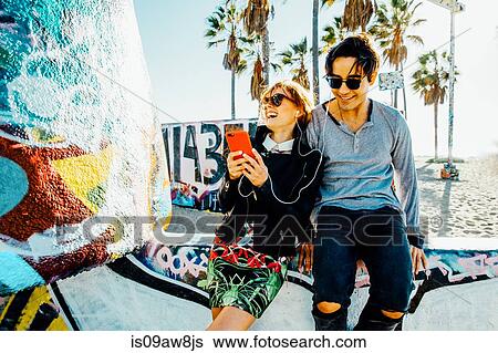 Stock Photograph - Young couple sitting on wall, sharing earphones, laughing. Fotosearch