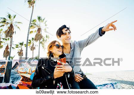 Young couple sitting on wall, sharing earphones, looking into distance View Large Photo Image Stock Photograph - Young couple sitting on wall, sharing earphones, looking into distance. Fotosearch