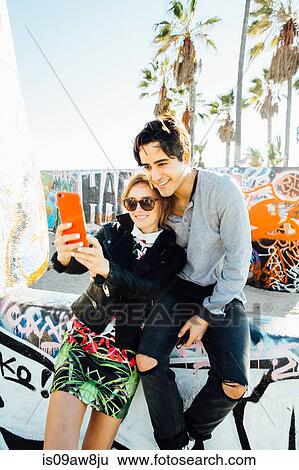 Stock Photograph - Young couple sitting on wall, taking self portrait using smartphone. Fotosearch