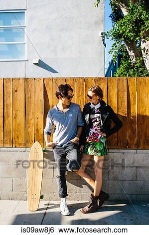 Young couple standing together outdoors, skateboard leaning on wall next to man View Large Photo Image Stock Photograph - Young couple standing together outdoors, skateboard leaning on wall next to man. Fotosearch