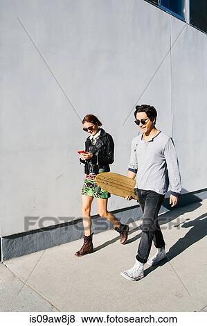Stock Photo - Young couple walking together outdoors, woman using smartphone, man carrying skateboard. Fotosearch