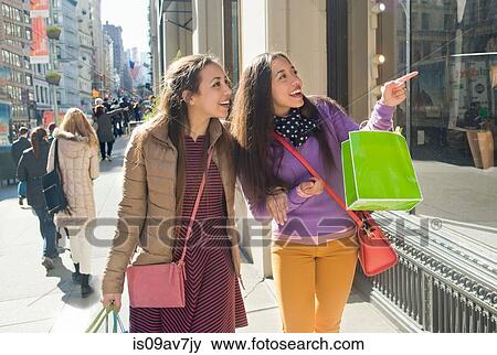 Stock Photo - Young female adult twins carrying shopping bags pointing at city shop window. Fotosearch