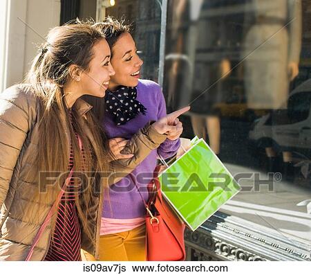 Stock Photograph - Young female adult twins carrying shopping bags pointing at city shop. Fotosearch
