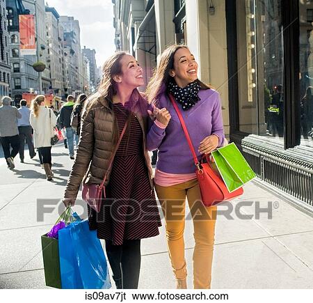 Stock Photograph - Young female adult twins carrying shopping bags strolling and chatting on city sidewalk. Fotosearch
