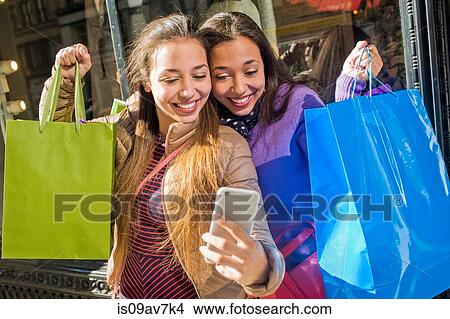 Young female adult twins in city taking smartphone selfie with shopping bags View Large Photo Image Picture - Young female adult twins in city taking smartphone selfie with shopping bags. Fotosearch
