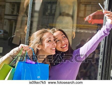 Stock Photography - Young female adult twins in city taking selfie on smartphone with shopping bags. Fotosearch