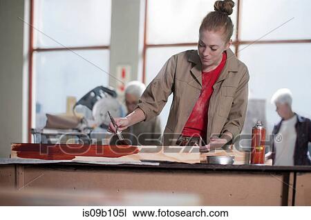 Stock Image - Young female carpenter painting wood stain in furniture making workshop. Fotosearch